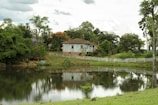 A rustic house with a blue and white exterior sits on a slight elevation surrounded by lush greenery and trees. A serene pond reflects the house and foliage, while a white picket fence lines the perimeter of the property. The sky is overcast with clouds, adding to the tranquil countryside setting.