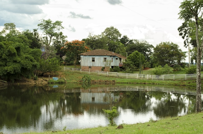 A rustic house with a blue and white exterior sits on a slight elevation surrounded by lush greenery and trees. A serene pond reflects the house and foliage, while a white picket fence lines the perimeter of the property. The sky is overcast with clouds, adding to the tranquil countryside setting.