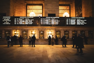 A busy ticket counter in a train station with people lined up to purchase tickets. The station is illuminated with warm lighting from large chandeliers above. Several screens display departure information for different train lines on a large board.