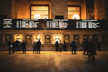 A busy ticket counter in a train station with people lined up to purchase tickets. The station is illuminated with warm lighting from large chandeliers above. Several screens display departure information for different train lines on a large board.