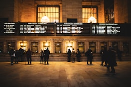 A busy ticket counter in a train station with people lined up to purchase tickets. The station is illuminated with warm lighting from large chandeliers above. Several screens display departure information for different train lines on a large board.