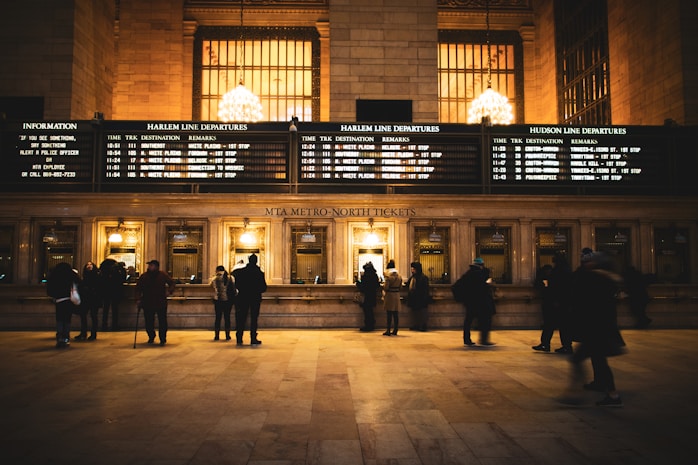 A busy ticket counter in a train station with people lined up to purchase tickets. The station is illuminated with warm lighting from large chandeliers above. Several screens display departure information for different train lines on a large board.