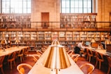 A spacious library features high bookshelves filled with a variety of books. There are wooden tables with brass reading lamps arranged in rows. One person is seen sitting at a table working on a computer. The room has large windows that allow natural light to flood in, adding a warm and inviting atmosphere.