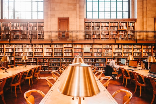 A spacious library features high bookshelves filled with a variety of books. There are wooden tables with brass reading lamps arranged in rows. One person is seen sitting at a table working on a computer. The room has large windows that allow natural light to flood in, adding a warm and inviting atmosphere.