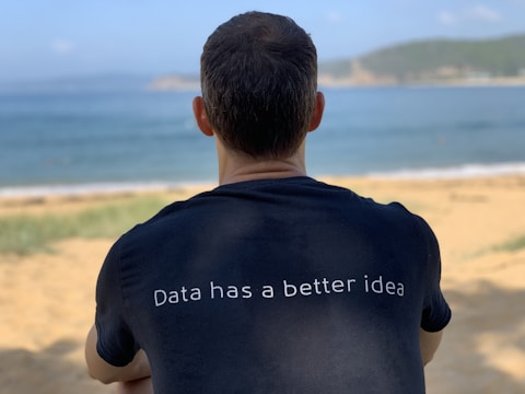 A person with short hair is sitting on a sandy beach, facing the ocean. They are wearing a dark t-shirt with the phrase 'Data has a better idea' printed on the back. The background includes blue water and distant hills under a clear sky.