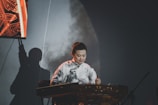 A person is playing a traditional stringed musical instrument, possibly a guzheng or a similar instrument, on a stage. They are wearing a patterned white and black top, and there is a shadow of the person visible on the backdrop behind them. There is an illuminated section on the left side of the image, displaying an abstract pattern.