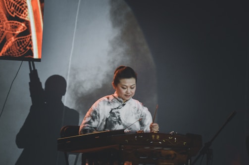 A person is playing a traditional stringed musical instrument, possibly a guzheng or a similar instrument, on a stage. They are wearing a patterned white and black top, and there is a shadow of the person visible on the backdrop behind them. There is an illuminated section on the left side of the image, displaying an abstract pattern.