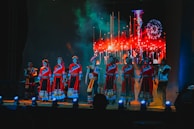 A group of performers stands on a stage dressed in traditional vibrant red attire with intricate patterns. The background features a bright, colorful display simulating fireworks or a festival scene. The stage lighting in blue tones highlights the performers, creating a dynamic atmosphere.