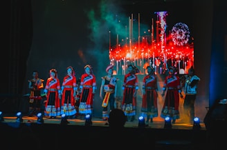 A group of performers stands on a stage dressed in traditional vibrant red attire with intricate patterns. The background features a bright, colorful display simulating fireworks or a festival scene. The stage lighting in blue tones highlights the performers, creating a dynamic atmosphere.