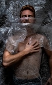 A person is lying down covered in clear bubble wrap. The hand is placed on the chest, and the face is partially obscured by the plastic material. The lighting creates dramatic shadows on the body and bubble wrap, emphasizing texture.