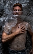 A person is lying down covered in clear bubble wrap. The hand is placed on the chest, and the face is partially obscured by the plastic material. The lighting creates dramatic shadows on the body and bubble wrap, emphasizing texture.