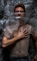 A person is lying down covered in clear bubble wrap. The hand is placed on the chest, and the face is partially obscured by the plastic material. The lighting creates dramatic shadows on the body and bubble wrap, emphasizing texture.