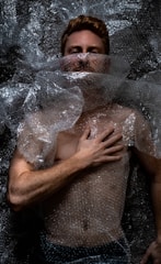 A person is lying down covered in clear bubble wrap. The hand is placed on the chest, and the face is partially obscured by the plastic material. The lighting creates dramatic shadows on the body and bubble wrap, emphasizing texture.