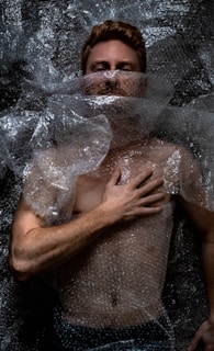 A person is lying down covered in clear bubble wrap. The hand is placed on the chest, and the face is partially obscured by the plastic material. The lighting creates dramatic shadows on the body and bubble wrap, emphasizing texture.