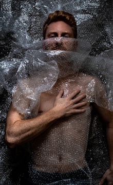 A person is lying down covered in clear bubble wrap. The hand is placed on the chest, and the face is partially obscured by the plastic material. The lighting creates dramatic shadows on the body and bubble wrap, emphasizing texture.