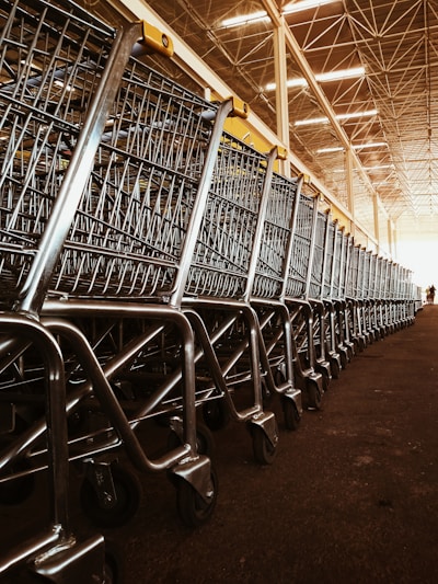 A large group of shopping carts arranged in a neat, orderly line inside a spacious warehouse. The metallic frames of the carts reflect the overhead lighting, and the ceiling features a grid-like structure with bright lights.