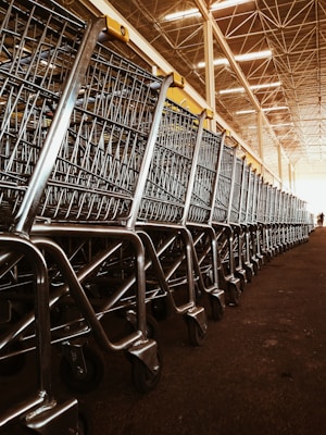 A large group of shopping carts arranged in a neat, orderly line inside a spacious warehouse. The metallic frames of the carts reflect the overhead lighting, and the ceiling features a grid-like structure with bright lights.