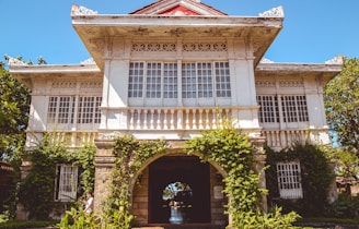 A large traditional colonial-style house with intricate architectural details, featuring decorative elements on the roof and windows. The building is mostly white with some red accents on the roof. It is surrounded by greenery, including climbing plants and trees, enhancing its elegant and historical appearance.