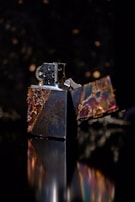 A metallic lighter with a reflective surface and intricate, colorful designs is captured against a dark, blurred background with bokeh lights. The open lid reveals the flame mechanism, which includes a windproof feature.