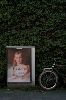 A poster advertising an art exhibition is placed against a lush green ivy-covered wall. The poster features a portrait of a young girl with a serene expression, announcing an exhibition at the Museum Van Loon from March 8 to June 4. A bicycle is positioned next to the poster, partially visible with its wheel and handlebars.