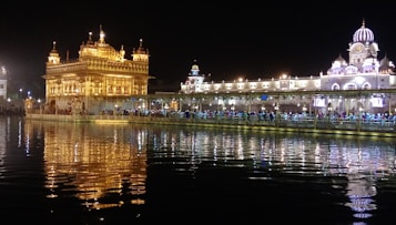 A majestic golden temple illuminated at night, reflecting off a serene body of water. The surrounding structures are also lit with vibrant lights, creating a beautiful and tranquil atmosphere.