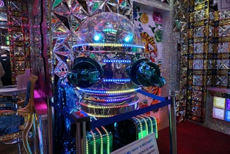 A highly reflective robot with a spherical head and glowing blue and green lights. The environment features colorful and intricate patterns with mirrored surfaces and geometric designs. There is a sign with text in front of the robot, and the walls display colorful, cartoonish tiger illustrations.