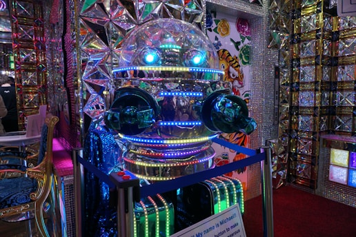 A highly reflective robot with a spherical head and glowing blue and green lights. The environment features colorful and intricate patterns with mirrored surfaces and geometric designs. There is a sign with text in front of the robot, and the walls display colorful, cartoonish tiger illustrations.