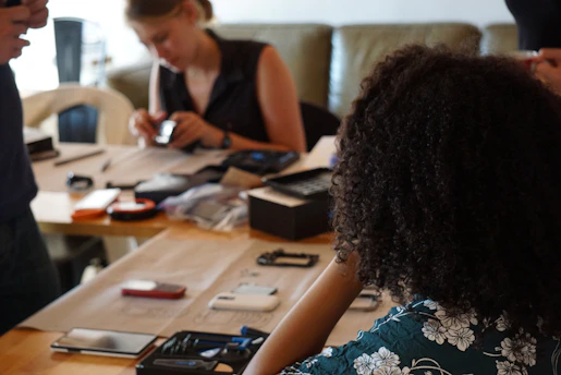 A group of people are gathered around a wooden table, which is strewn with various electronic devices and components. The focus is on a person with curly hair in the foreground, who is looking at the table. Another individual is sitting at the table, examining a device in their hands, while more people and gadgets are visible in the background.