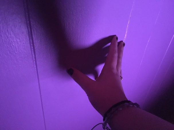 A hand with painted nails extends towards a vertical surface illuminated in purple light. The hand casts a shadow on the wall, creating a striking visual effect.