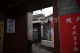 An alleyway with traditional Chinese architecture featuring a narrow path lined with signs displaying Chinese characters and a small window display. There are several red signs and banners, adding a splash of color against the gray stone walls. The atmosphere feels authentic and reminiscent of old Beijing.