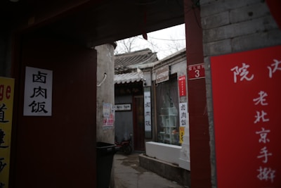 An alleyway with traditional Chinese architecture featuring a narrow path lined with signs displaying Chinese characters and a small window display. There are several red signs and banners, adding a splash of color against the gray stone walls. The atmosphere feels authentic and reminiscent of old Beijing.