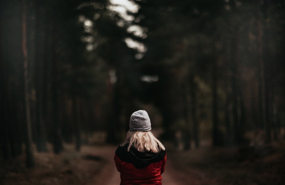 woman wearing grey beanie in red hoodie standing on pathway between green leafed trees during daytime