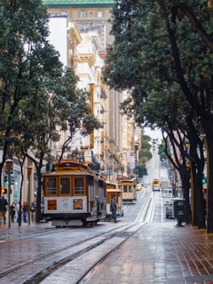 A bustling urban street scene features iconic historic streetcars traveling along tracks lined with trees, buildings, and people walking on the sidewalks. The view stretches up a steep hill with a mix of architectural styles visible, making for a vibrant and dynamic city atmosphere.