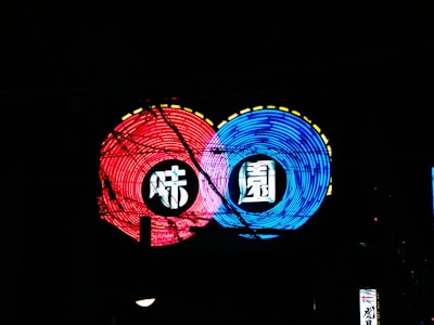 A vibrant neon sign with two circular designs, one in red and the other in blue. Each circle contains a stylized symbol or character. Yellow dashes outline parts of the circles while black cables or wires intersect the neon lights, adding an urban feel.
