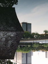 An inverted image featuring a traditional stone building on the left with a large, modern office building in the background. A serene body of water reflects the buildings and surrounding trees, creating a tranquil setting.
