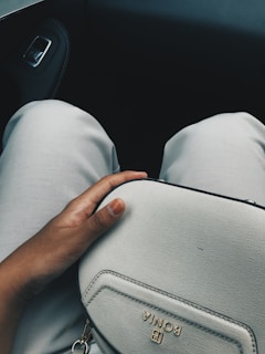 A hand rests on a light-colored, textured handbag with the brand name 'BONIA' embossed in gold. The person is seated in a vehicle, indicated by the visible car door and seat legs in the background. The legs are dressed in light-colored pants, and the lighting suggests a calm and relaxed setting.