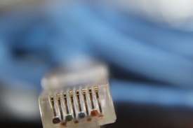 A close-up view of an Ethernet cable connector, highlighting its metallic pins. The background is blurry, featuring a coiled blue cable.