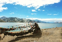 A serene lakeside landscape with vibrant prayer flags strung across a rocky shore. The backdrop features majestic mountains under a bright blue sky with scattered clouds. The water reflects the colors of the sky and surrounding environment.