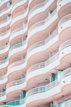 A close-up view of a modern apartment building featuring multiple levels of curved balconies. The balconies have white railings and are complemented by accents of soft pastel colors, including light pink and turquoise. There are glass doors leading to each balcony, some of which are open.