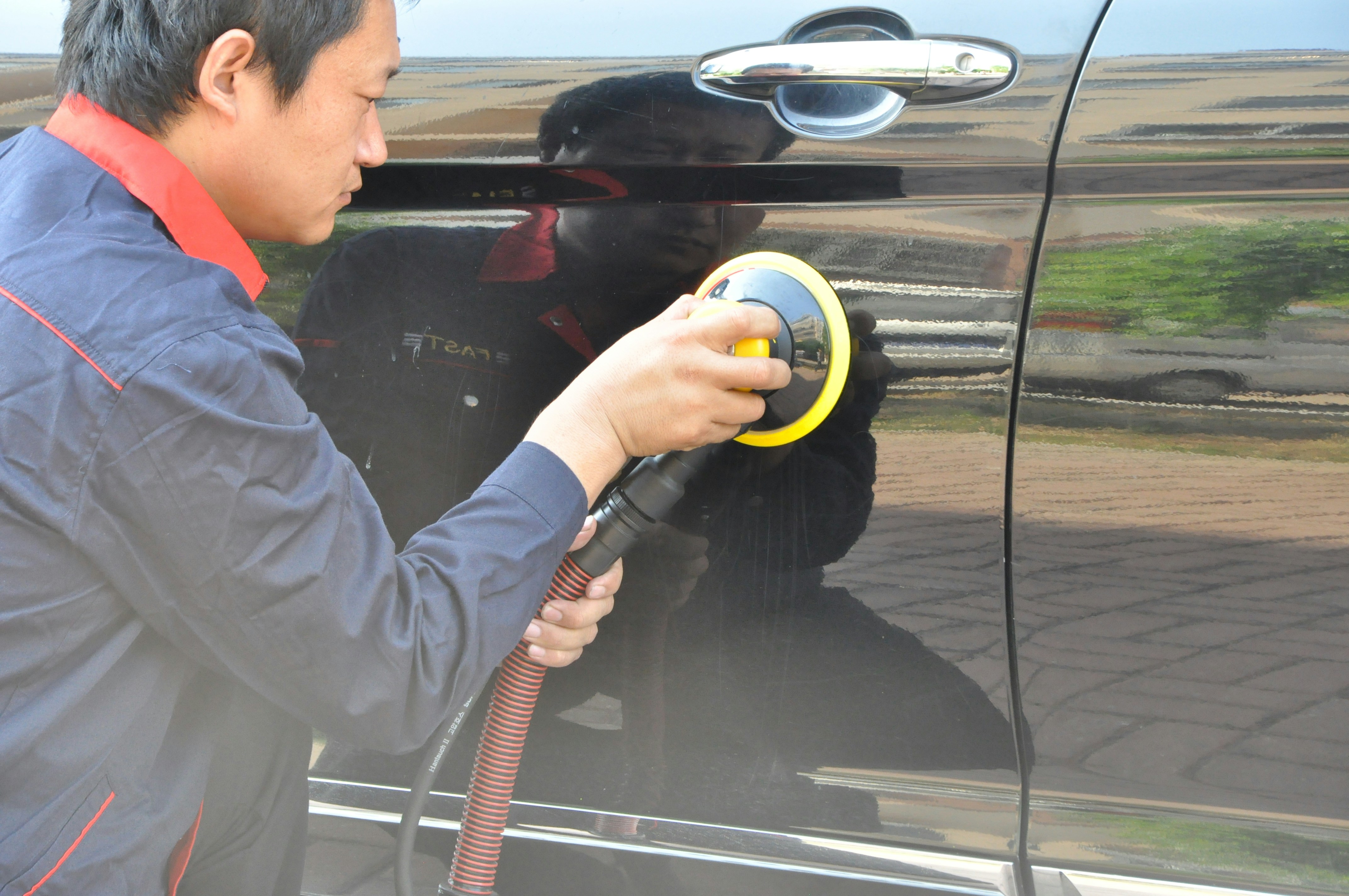 A person is using a polishing tool on the side of a black car, focusing on applying an even pressure and achieving a smooth finish. The reflection on the car surface shows the surrounding environment.