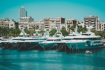 Several luxurious yachts are moored in a marina adjacent to a cityscape featuring multi-story buildings. The water in the foreground is calm and reflects the yachts and the clear sky.