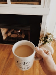 A hand holds a white mug filled with coffee in front of a cozy fireplace. The mug has a handwritten-style text design on it. The fireplace is built into a wall, with natural logs visible inside and a gentle flame burning. The wooden floor adds warmth to the setting, and a subtle floral arrangement can be seen to the side.