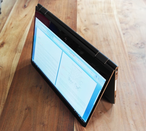 A convertible laptop is positioned in a tent mode on a wooden table, displaying a document on its screen. The table has a smooth finish with visible grains, and the laptop appears to be slim and modern.