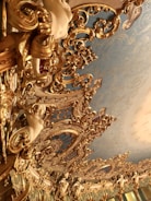 An ornate and intricately designed ceiling features elaborate gold gilded patterns and decorative sculptures of cherubs and other figures against a backdrop of soft blue paint. The ceiling is highly detailed, with swirling motifs and rich textures.