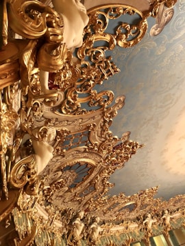 An ornate and intricately designed ceiling features elaborate gold gilded patterns and decorative sculptures of cherubs and other figures against a backdrop of soft blue paint. The ceiling is highly detailed, with swirling motifs and rich textures.