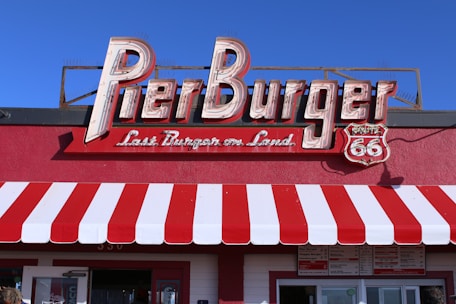 A retro-style restaurant sign with neon letters spells out 'Pier Burger'. Below the sign, there is text stating 'Last Burger on Land' next to a Route 66 emblem. The building facade features a bold red and white striped awning.