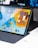 A tablet with a vibrant, colorful display shows various app icons on its screen, positioned in a keyboard dock setup. The screen features an orange, blue, and yellow background with app widgets on the right-hand side. A dark background monitor is visible in the rear, partially displaying its own contents. Alongside, there's a wireless earbud case on the desk.