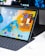 A tablet with a vibrant, colorful display shows various app icons on its screen, positioned in a keyboard dock setup. The screen features an orange, blue, and yellow background with app widgets on the right-hand side. A dark background monitor is visible in the rear, partially displaying its own contents. Alongside, there's a wireless earbud case on the desk.