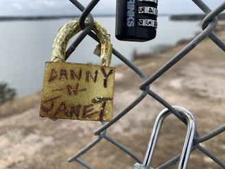 A close-up of a chain-link fence with two locks attached. One lock is golden and has names written on it, while the other is a black combination lock. The background includes a hazy view of a body of water and shoreline, suggesting a secluded or tranquil setting.