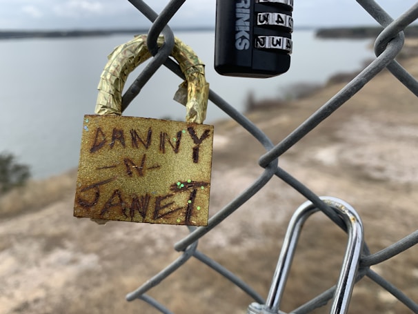 A close-up of a chain-link fence with two locks attached. One lock is golden and has names written on it, while the other is a black combination lock. The background includes a hazy view of a body of water and shoreline, suggesting a secluded or tranquil setting.